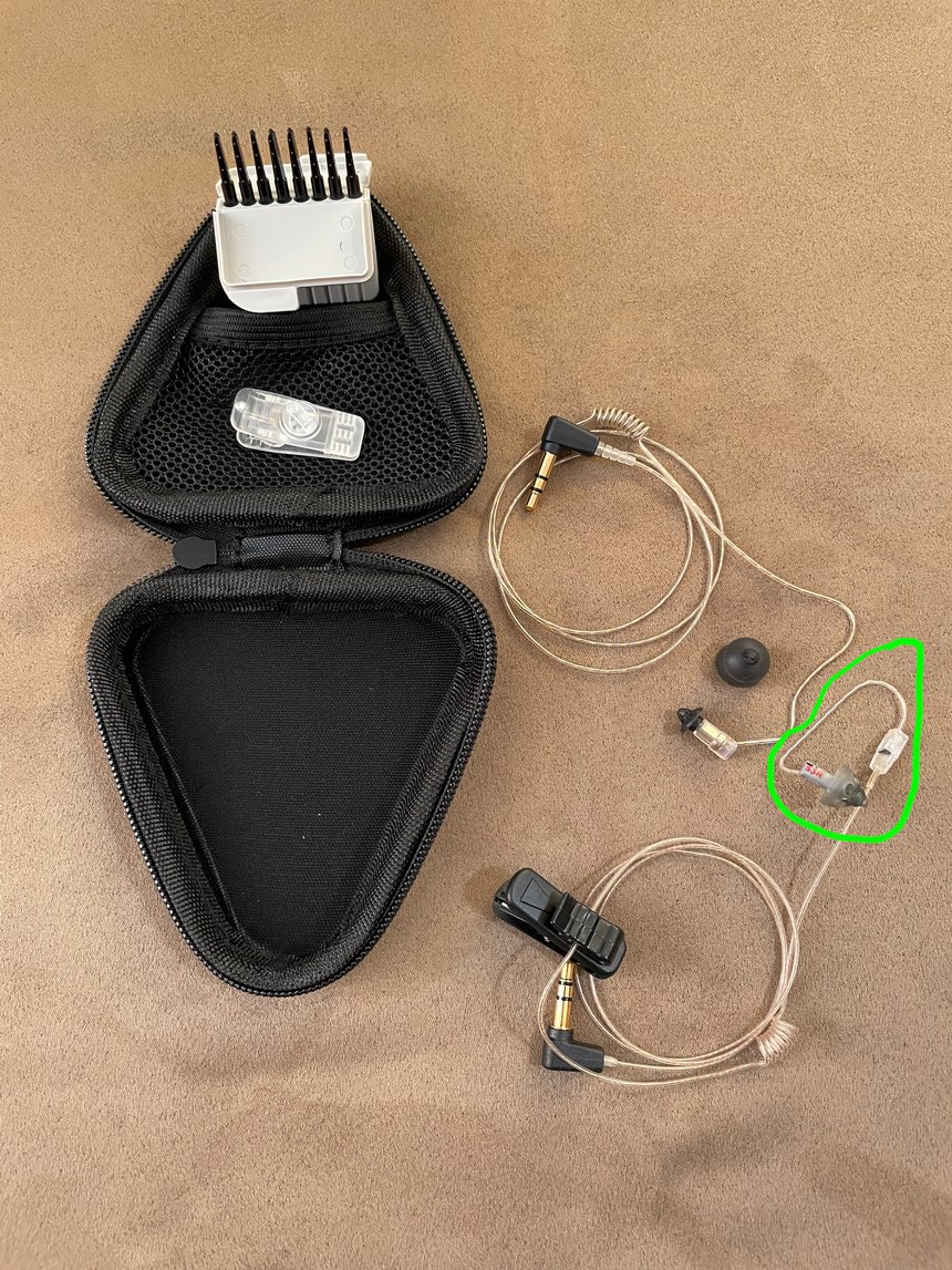 Clockwise from left top: N•ear case holding ear wax remover tools and a collar clip, the new softer-wire earpiece and the first-gen earpiece. Note the first-gen’s stiff wire and junction box, which connects the hearing aid speaker to their wire.