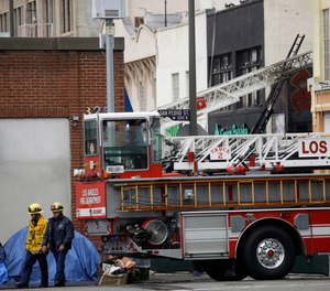 Firefighters walk the scene of a fire and explosion at a smoke and vape wholesale business on Monday, May 18, 2020, in Los Angeles. The Fire Department was at the nondescript commercial building late Saturday afternoon when there was a thunderous explosion that sent firefighters scrambling through searing flames to get out of the building. The department now plans to conduct a citywide review to examine how similar businesses store volatile materials.