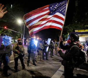 A demonstrator waves a U.S. flag in front of federal officers after tear gas is deployed during a protest at the Mark O. Hatfield United States Courthouse Thursday, July 30, 2020, in Portland, Ore. (AP Photo/Marcio Jose Sanchez)