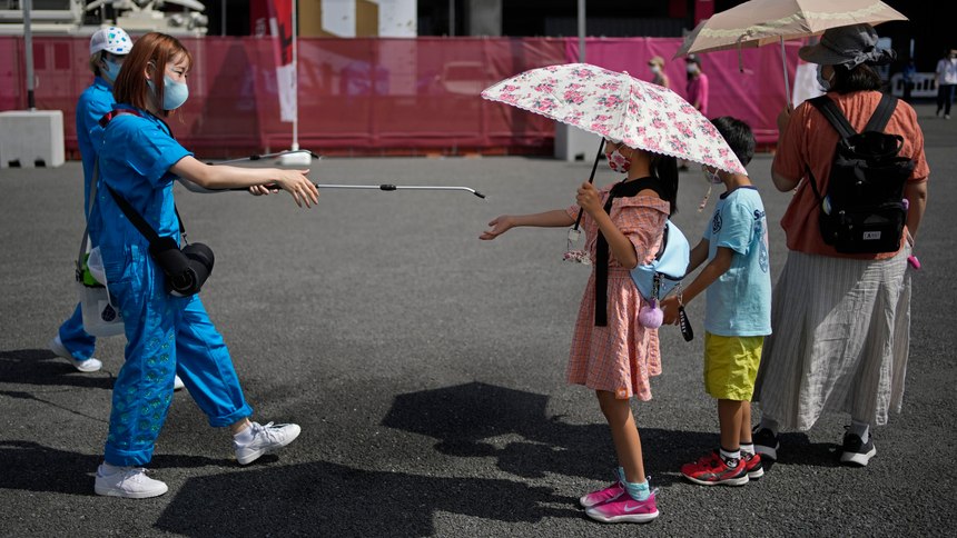 A volunteer sprays water on children as they use umbrellas to beat the heat outside the Fuji International Speedway, the finish for the women's cycling road race that is underway, at the 2020 Summer Olympics, Sunday, July 25, 2021, in Oyama, Japan.