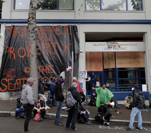 In this photo taken June 11, 2020, people sit in front of the Seattle Police Dept.'s East Precinct headquarters, which at the time was boarded up and abandoned except for a few officers inside in an area of Seattle that was occupied when demonstrators took over part of the city's Capitol Hill neighborhood.