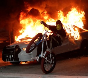 A protester on a bicycle rides past a burning police car during a protest next to the city of Miami Police Department, Saturday, May 30, 2020, downtown in Miami.
