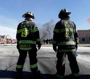 Firefighters waiting to load load their truck with water look on as a news conference is held in front of smoke rising at the site of a large warehouse fire a day after flames engulfed the facility, Friday, Feb. 12, 2016, in Hillsborough, N.J.