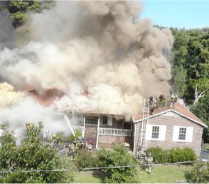 A drone films the exterior of a house fire, watching crews operating on roof.