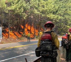 Sourdough Fire crews stand along the opposite edge of State Route 20 as other firefighters apply water from hoses supplied by a water tender truck.