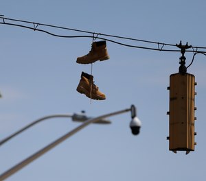 In this Wednesday, April 29, 2020 photo, a pair of work boots hang from the wire of a traffic light with a surveillance camera seen in the distance in West Baltimore.