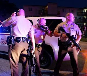 Police officers stand near a crowd that is gathering by the scene where 18-year-old Michael Brown was fatally shot by police in Ferguson, Mo., near St. Louis on Saturday, Aug. 9, 2014.