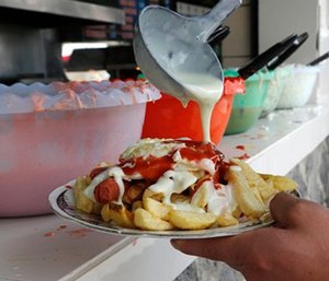 A man pours mayonnaise over his plate of french fries and sausage at a fast food stand.