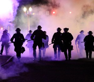 In this March 29, 2020, file photo, police officers walk enveloped by teargas in Portland, Ore., a liberal city with a reputation for full-throated and frequent protests.