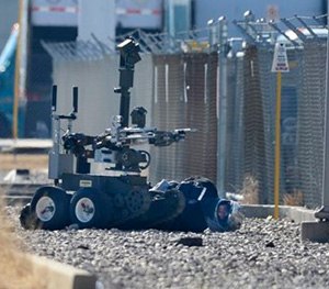 A bank robbery suspect wearing a motorcycle helmet lies handcuffed on the ground as a bomb squad robot searches his vest.