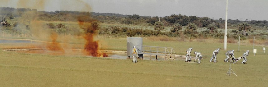 Astronaut Rescue team practices rescue drill at the Hypergolic Fire Training facility. Nitrogen tetroxide and monomethylhydrazine is released from storage tanks (not shown) and ignite when the products flow together in a pan to the left of the Mock Command Module (center). The Astronaut Rescue Team extracts fill-in Astronaut from Mock command module.